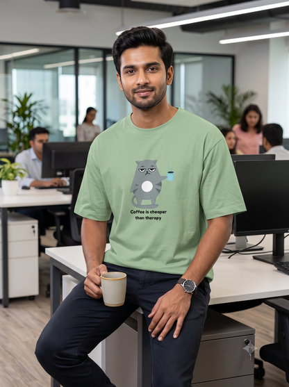 A man in an office setting wearing a light green t-shirt with a cartoon cat graphic and the text 'Coffee is cheaper than therapy'. 
