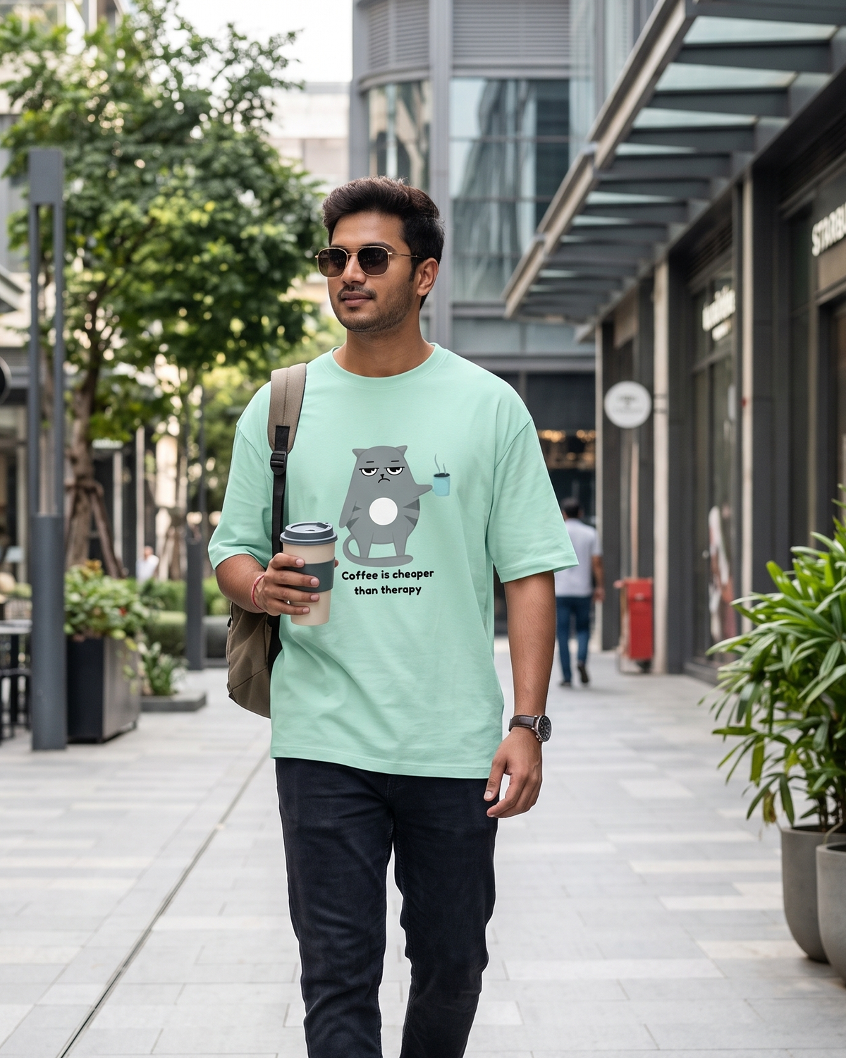 A man walking on a modern outdoor shopping plaza wearing a light mint green graphic t-shirt with a cartoon cat and the text 'Coffee is cheaper than Therapy'