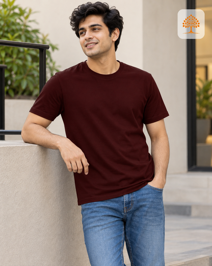 A man wearing a dark maroon crew-neck t-shirt and blue jeans, leaning against a concrete wall in an outdoor setting. A small logo is visible in the top right corner.