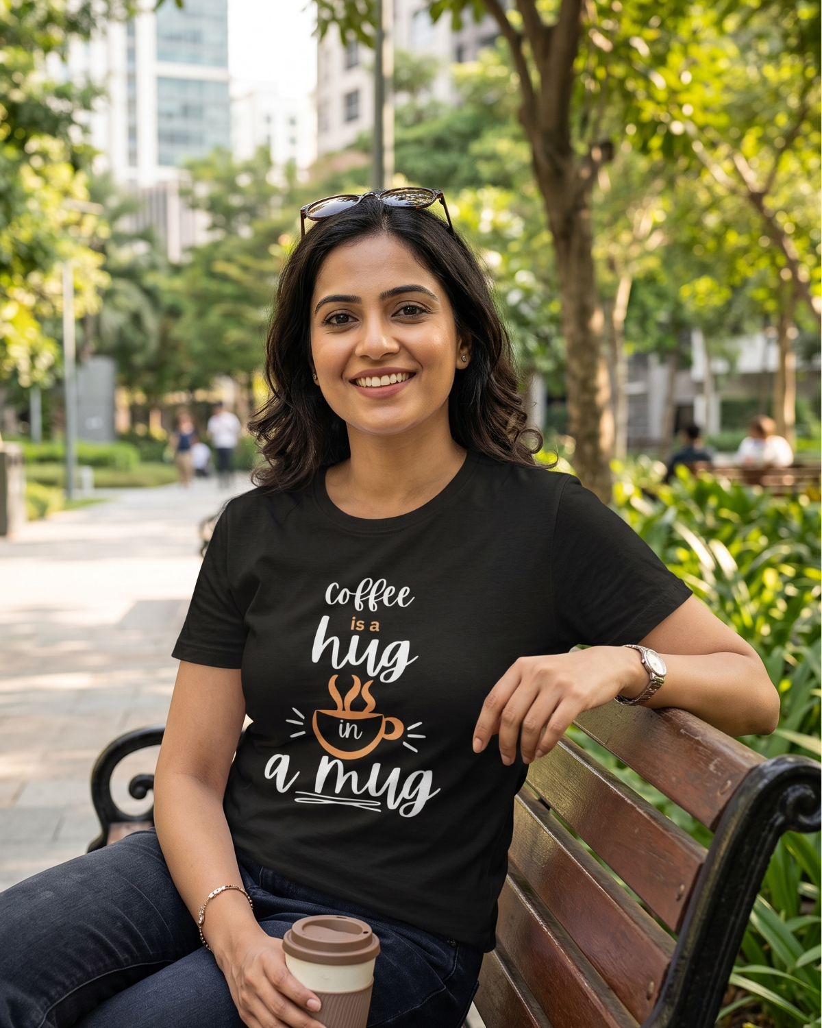 A woman sitting on a park bench wearing a black graphic t-shirt with the text 'coffee is a hug if it's a mug' in white and orange. She is holding a paper coffee cup, with a blurred green park background.